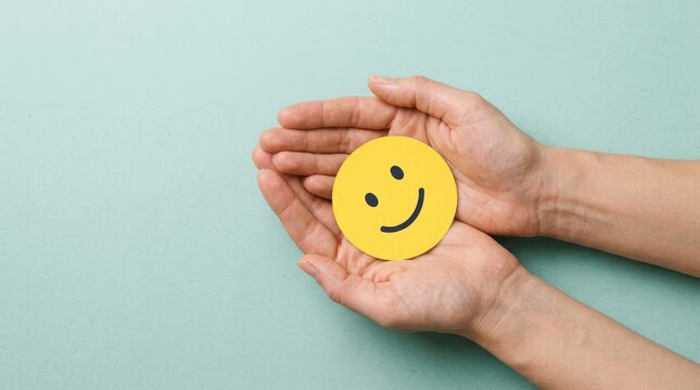 Two open human hands gently cradling a bright yellow paper circle featuring a simple black hand drawn smiling face icon