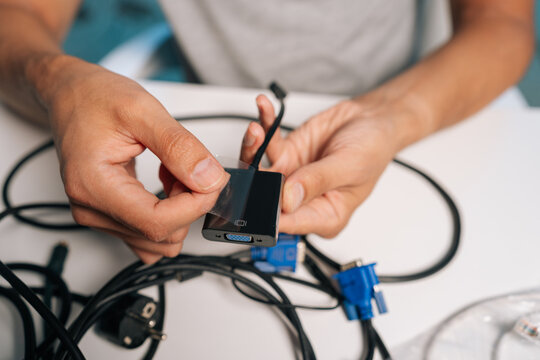 Close-up hands of unrecognizable person peeling transparent protective film from new black video adapter, preparing device for connection amidst tangle of cables and connectors on white desk.