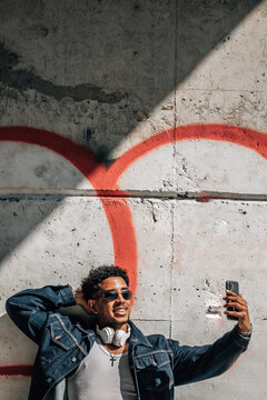 Young african american man smiling confidently while taking a selfie with his smartphone against an urban graffiti wall, expressing joy and lifestyle energy in an outdoor street