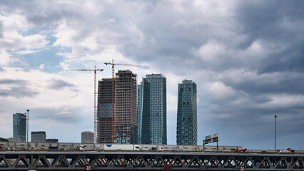 Tall high rise buildings and cranes rise behind a busy bridge. Dark storm clouds frame the growing city skyline. Urban development and infrastructure concept. © jahorimine