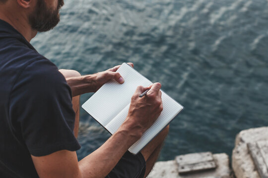 Close-up of man writing in notebook while sitting on pier above the water during a quiet summer evening, concept of creativity and solitude