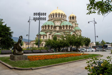 Summer view of Sofia, Bulgaria, featuring the St. Alexander Nevsky Cathedral with its golden and green domes under a dramatic cloudy sky, with a bronze sculpture, flower beds and greenery in the foreg © snatalia