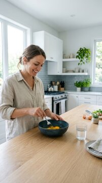 Smiling woman whisks eggs in a deep bowl on a bright, sunlit kitchen island, perfect for lifestyle advertising with copy space.