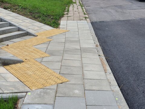 Outdoor urban infrastructure scene showing a paved sidewalk with light grey rectangular stone tiles adjacent to an asphalt road. Prominent yellow tactile paving tiles with raised dome patterns are