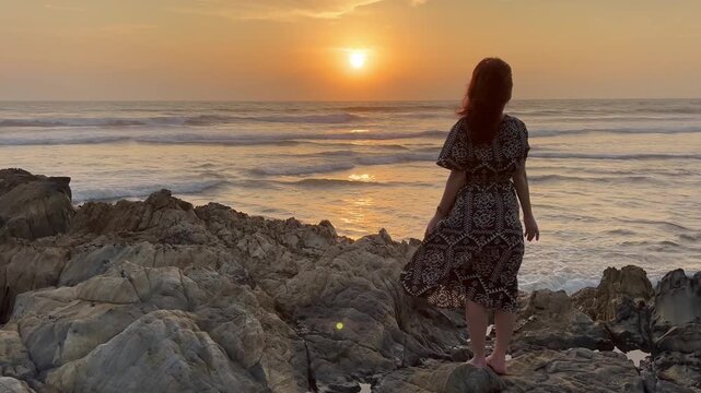 Static shot of woman with red hair standing on coastal rocks with open palms, meditating during a golden ocean sunset