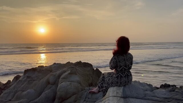 Static shot of lonely woman with red hair sitting on coastal rocks, feeling sad and thoughtful during ocean sunset