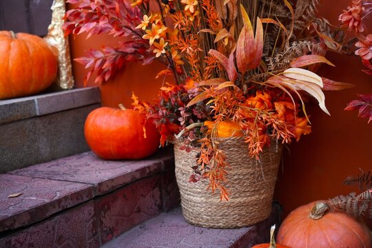 Orange pumpkins and autumn flowers arrangement in woven basket decorating home porch steps for fall harvest festival and Halloween. Seasonal porch decor with warm orange color palette.