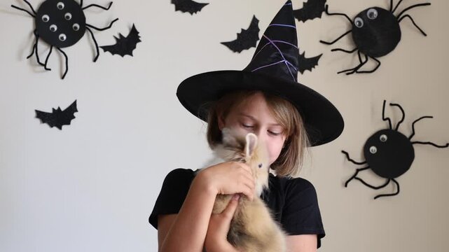 Video portrait happy excited emotional face girl child in black witch hat holding orange candy bucket rabbit against wall decorated background bats. handmade decorations. Pet shop advertising