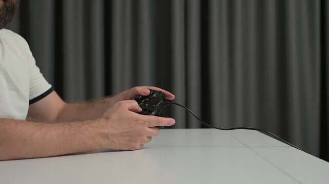 Close up hands holding wired black game controller over glossy white tabletop, thumbs poised above buttons, cable trailing toward gray curtain backdrop in indoor studio