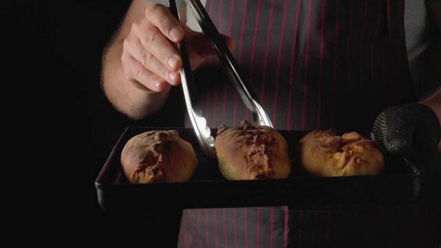 Baker in striped apron uses tongs to lift freshly baked bread rolls from a tray, showcasing the golden crust and soft texture against a dark background in a kitchen setting
