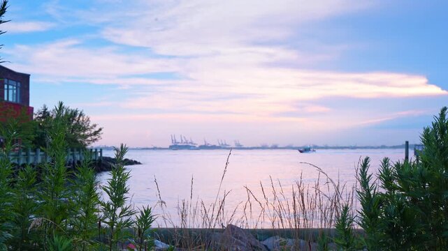 River landscape with coastal vegetation and distant port cranes in New York. A small boat navigates calm waters framed by green plants under a soft pastel evening sky.