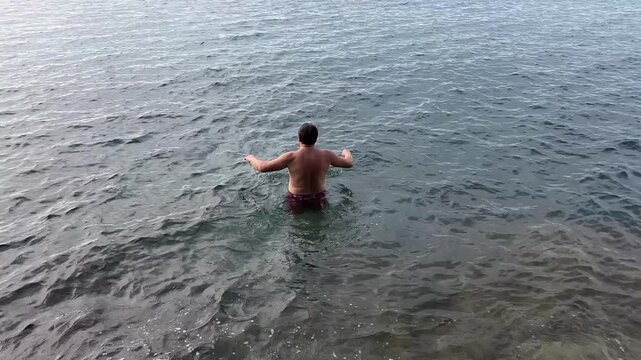 Adult man standing waist deep in sea water spreading arms and diving headfirst into waves during sunny summer vacation day.