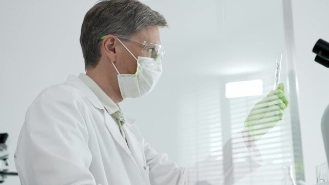 Male scientist wearing a white lab coat, safety glasses, mask and green gloves, analyzing a small plant sample in a test tube while conducting a botanical experiment in a clean laboratory