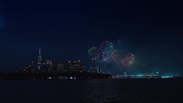 New York night skyline with green and red firework bursts.. Multicolored pyrotechnics explode above dark river water beside illuminated urban skyscrapers.