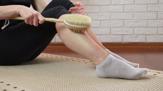 Woman performing dry brushing massage on her leg. Unrecognizable woman sitting on a floor mat while performing a dry brushing massage on her leg to reduce cellulite, improve circulation