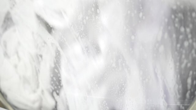 Fabric repeatedly passes behind a sudsy washing machine window during drum rotation, illustrating the wet processing stage of a mechanized laundry workflow