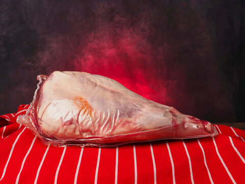 Raw lamb leg in plastic vacuum wrap rests on red and white table cloth, painted background. Meat shows pink flesh and red fat marbling. Scene suggests butchery, cooking, or grocery retail.