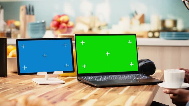 Young woman works indoors at a kitchen table with green screen laptop and blue screen tablet, multitasking with devices for screen replacement and mockup. Work from anywhere supports tasks at home.