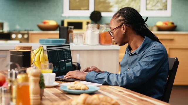 Woman transcribes data for research using laptop and digital tools at kitchen table. Telecommuting and outsourcing use charts and network connectivity for hybrid work while she works remotely.