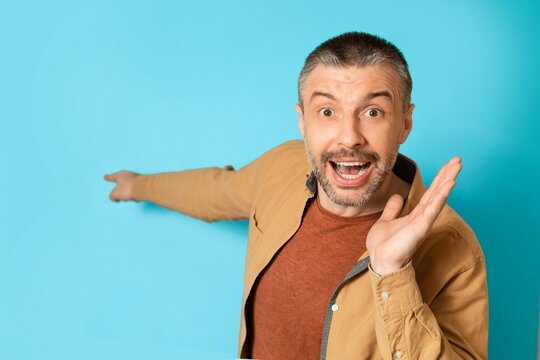 Excited Caucasian Man Smiling And Pointing At Copy Space, Standing In Front Of Solid Blue Background. Perfect For Business Concepts, Marketing Ads, Promotions, Or Advertisements.