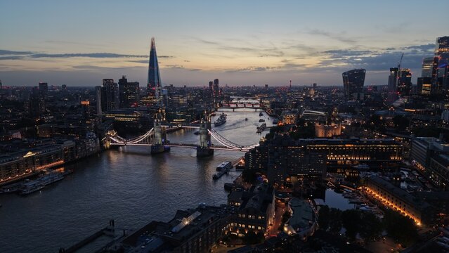 Aerial panoramic view of London at sunset with illuminated Tower Bridge crossing the River Thames, The Shard and the City of London skyline glowing in the evening.