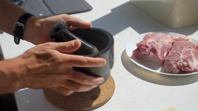 Man grinds spices with pestle in black mortar beside raw pork. Concept of preparing seasoning, cooking ingredients and home meal preparation