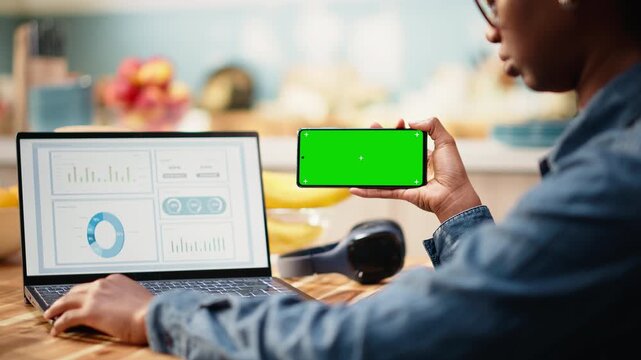 Isolated mockup on mobile phone screen is used at home in kitchen while woman remote worker analyzes metrics and charts on laptop. Teleworking and outsourcing support task management.