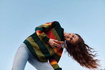 Plakat Young woman in a colorful striped sweater leans back laughing while holding her phone against a clear blue sky. Joyful carefree mood. Concept of happiness and freedom.