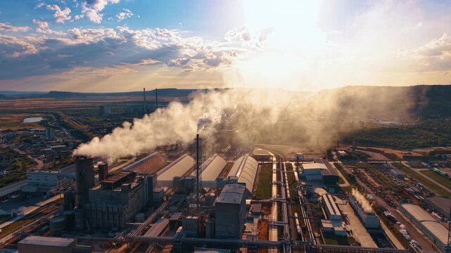 Heavy emissions fill the air above the factory buildings. The setting sun highlights the smog and structures.