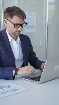 Professional businessman in a suit jacket and glasses focused on a laptop at his desk, reviewing documents and charts for finance and strategy in a modern office setting