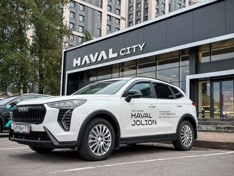 White Haval Jolion test drive SUV car parked outdoors on asphalt street next to a modern Haval City dealership showroom glass building facade. Premium Chinese crossover automobile transport vehicle. 
