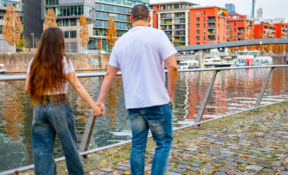 Happy young couple, happy girl and man stands together on waterfront promenade, modern apartment buildings across water, sharing quiet, intimate moment in contemporary urban landscape, cheerful mood