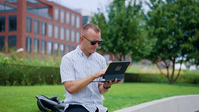 He wears casual clothes and sunglasses working near a backpack and brick architecture.