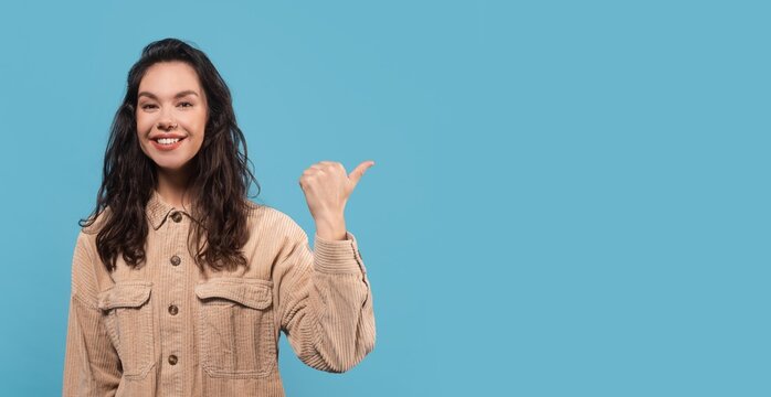Cheerful Young Woman Smiling And Pointing At Camera, Showcasing Positive Emotion In A Studio Setting With Blue Background. Engaging Expression Representing Happiness And Confidence.