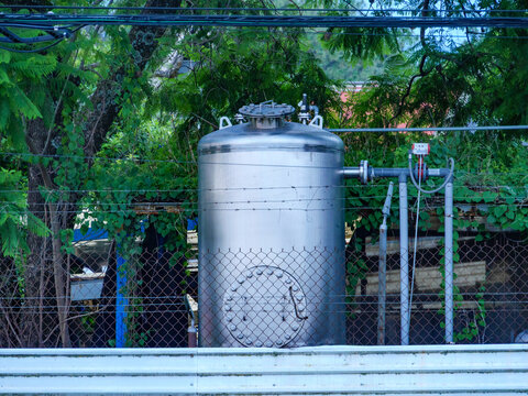 Gran tanque de almacenamiento industrial de acero inoxidable detr&aacute;s de una cerca de malla rodeado de follaje verde.