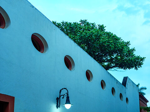 Detalle arquitect&oacute;nico abstracto de un muro blanco con ventanas circulares, un farol cl&aacute;sico encendido y la copa de un &aacute;rbol al atardecer.