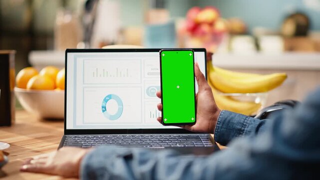 Vertical chroma key screen on smartphone used at home in kitchen, remote worker analyzing metrics and charts on laptop. Teleworking and outsourcing showing task management and research.
