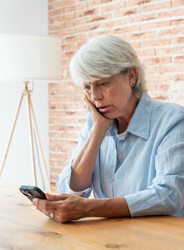 Concerned older woman sits at a table in a bright room, staring at her smartphone with a troubled expression. Natural light, casual clothing and exposed brick create an intimate, everyday scene.