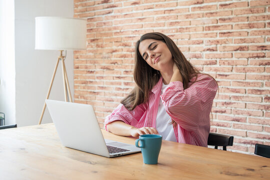 Young woman feeling neck pain and discomfort while working remotely on a laptop, indicating stress, fatigue, and stiffness from prolonged screen time and poor posture during remote work