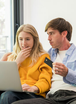 Young couple looking surprised while sitting on a couch, checking information on a laptop, and holding a credit card for online shopping or a financial transaction