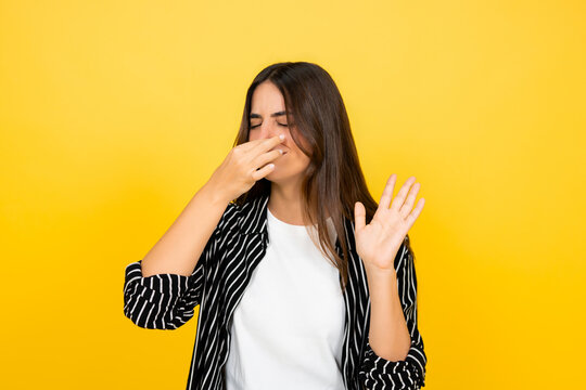 Young woman pinching her nose with a disgusted expression. Covering her face and waving off an unpleasant stink or odor. Showing repulsion and avoiding a foul smell against a vibrant yellow background