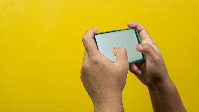 Two adult male hands grip a blank smartphone horizontally against a solid yellow background, demonstrating active mobile gaming or rapid text typing postures. Both thumbs rest naturally