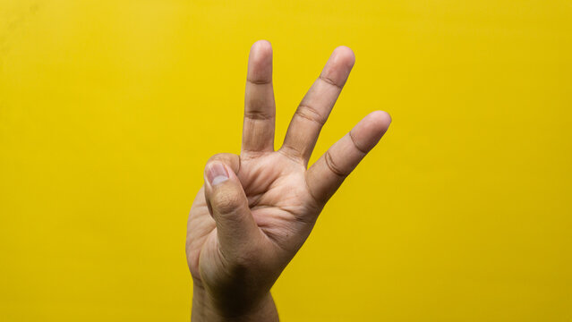 One male hand is raised indicating number three using extended digits against vivid yellow background, Index middle alongside ring fingers point straight upward while thumb securely