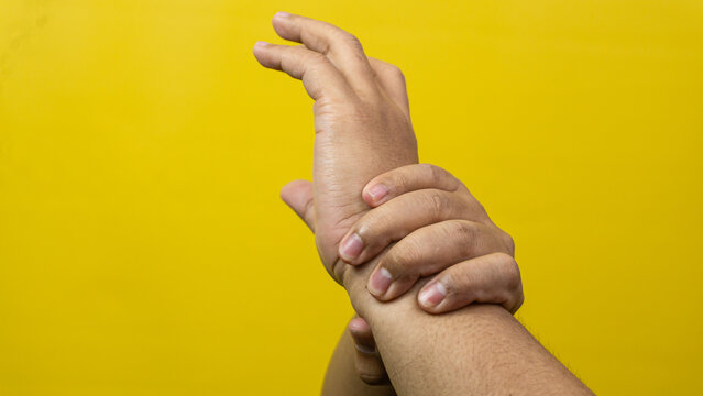 One adult male hand firmly grips the opposite wrist against a solid yellow background creating strong visual metaphors regarding restraint or medical support. Fingers wrap securely