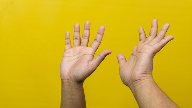 Two adult male hands are raised with open palms and spread fingers against a vibrant yellow background. This defensive or surrendering posture effectively communicates universal nonverbal