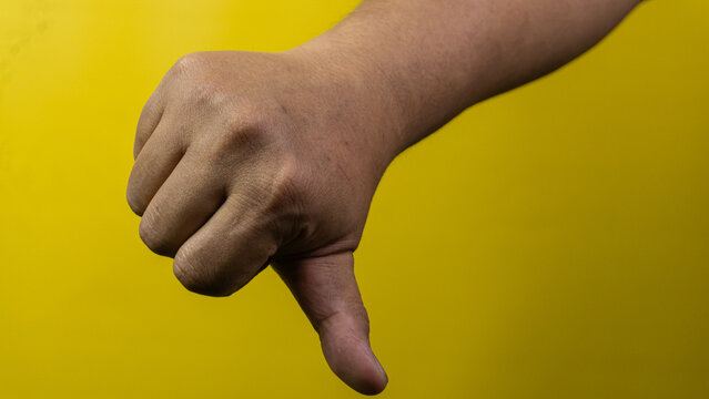 A right arm is captured showing a clear thumbs down gesture in front of a solid, bright yellow background. The hand is formed into a fist with the thumb pointing straight towards the