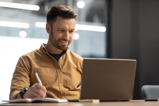 Focused Caucasian Man In Mid 30s Writing Notes And Using Laptop At Desk In Modern Office, Engaged In Work Tasks, Professional Environment, Business Concept.