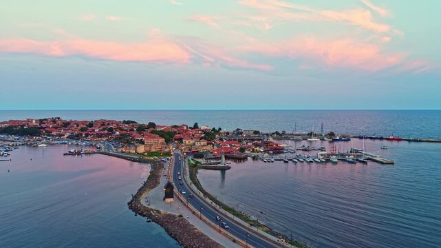 Aerial view of a coastal town with buildings along the shore, boats docked, and a road connecting to the harbor. The sky shows colors from the sunset.
