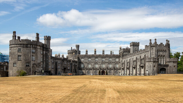 Historic Kilkenny Castle facade with stone towers and dry grass lawn under blue sky in Kilkenny, County Kilkenny, Ireland.