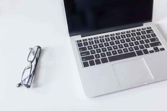 A sleek modern laptop and a pair of eyeglasses rest neatly on a plain white desk, forming a tidy workspace setup.